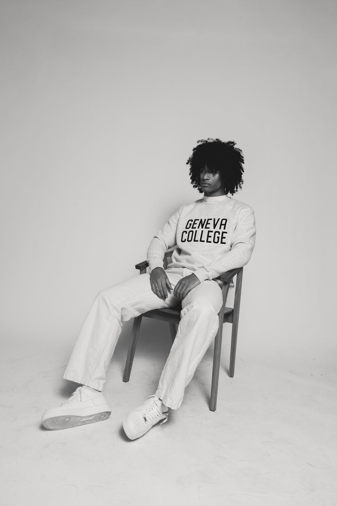 Male wearing a 'Geneva College' sweatshirt sitting on a chair against a plain background